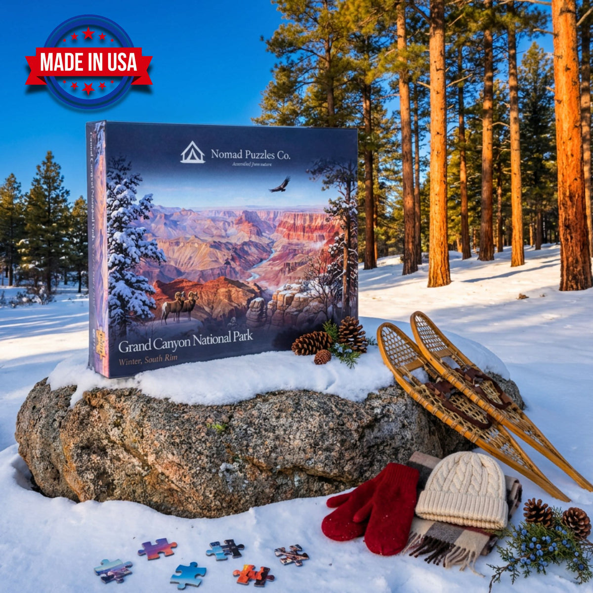 Nomad Puzzles Co. Grand Canyon Winter South Rim 1,000-piece puzzle box on a snow-dusted boulder in a ponderosa pine forest, with wooden snowshoes, red mittens, and a knit beanie