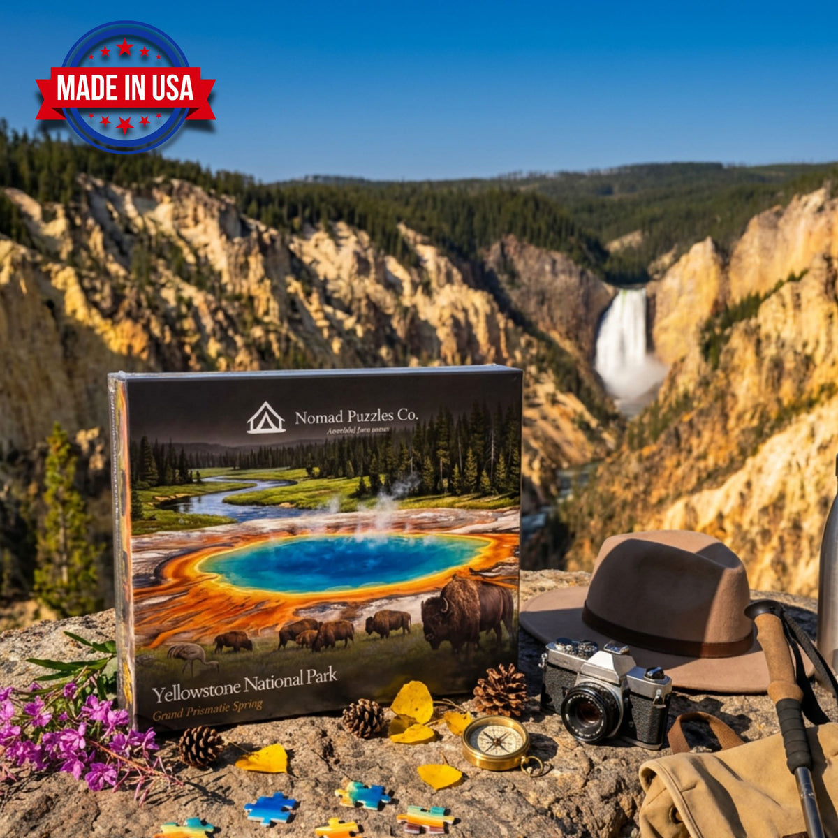 Nomad Puzzles Co. Yellowstone Grand Prismatic Spring 1,000-piece puzzle box on a rock ledge above the Grand Canyon of the Yellowstone and Lower Falls, with a felt hat, film camera, and compass