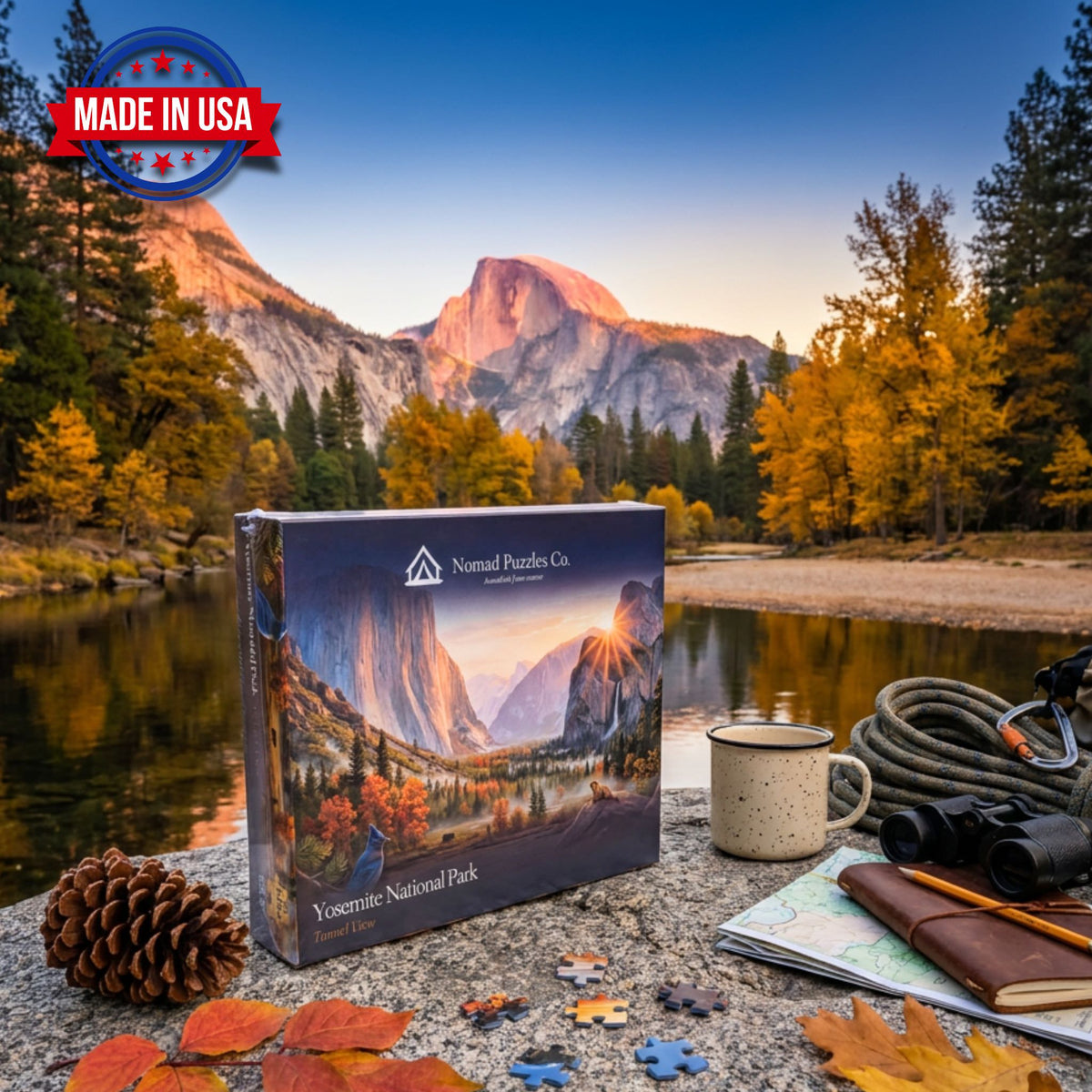 Nomad Puzzles Co. Yosemite Tunnel View 1,000-piece puzzle box on a granite boulder beside the Merced River, with Half Dome at sunset, autumn trees, and a pine cone