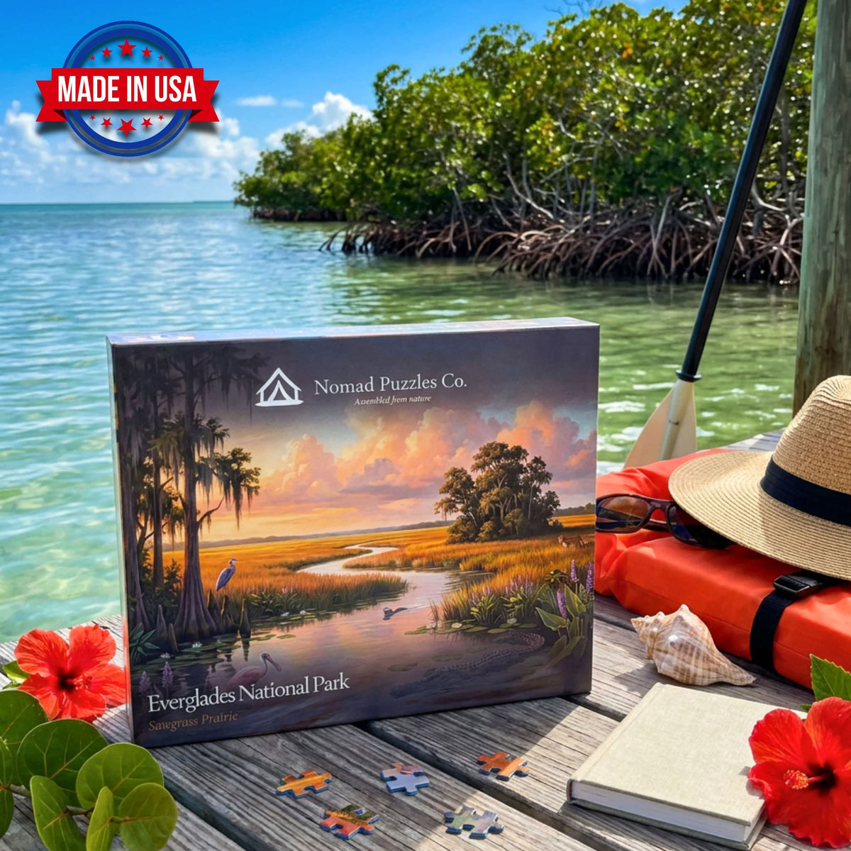 Nomad Puzzles Co. Everglades Sawgrass Prairie 1,000-piece puzzle box on a weathered dock beside turquoise Florida Bay, with mangroves, a straw hat, life vest, and kayak paddle